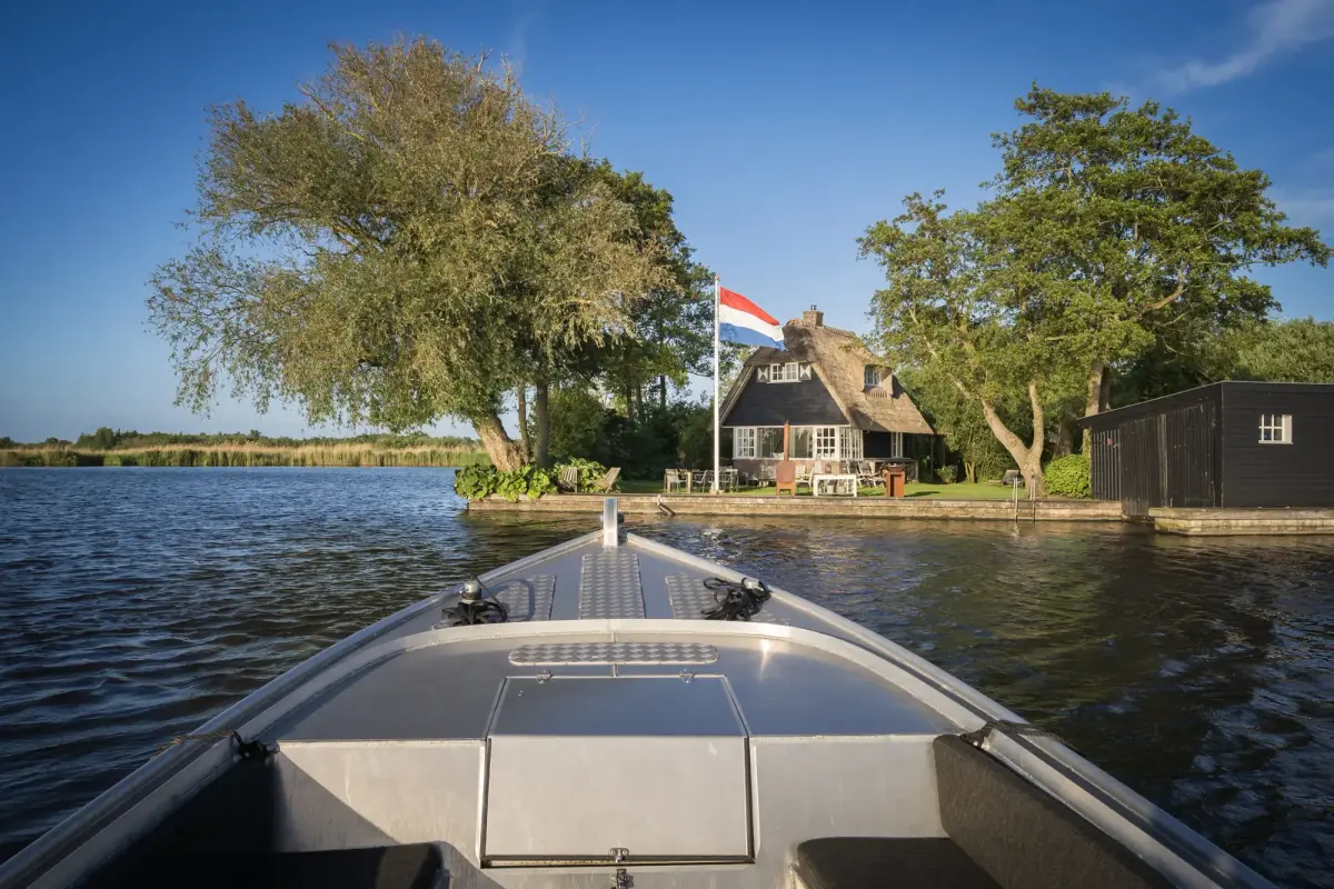 Vanaf de voorplecht van een sloep op weg naar een rietgedekt huis met een Nederlandse vlag aan de waterkant